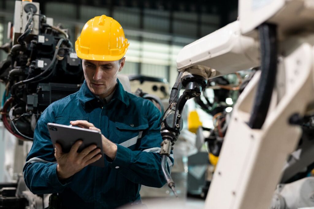 Mitarbeiter mit Schutzhelm bedient Industrieroboter über Tablet – moderne Automationstechnik im Produktionsumfeld